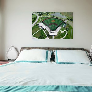 May include: An aerial photograph of a baseball field with green grass and a red infield. The field is surrounded by a white fence and green trees. The photo is hanging on a wall above a white bed with blue and white bedding.