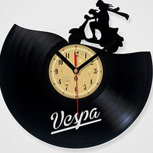 Könnte beinhalten: Schwarze Vespa-Uhr aus einer Schallplatte. Die Uhr zeigt eine Silhouette einer Person, die einen Roller fährt, und das Wort "Vespa" in weißer Schrift. Das Zifferblatt ist beige mit schwarzen Ziffern und Zeigern.