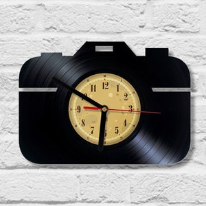 Puede incluir: Reloj de vinilo negro con forma de cámara. La esfera del reloj es de color crema con números y manecillas negros.