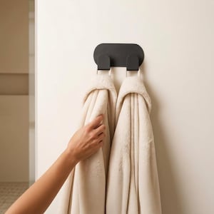 Pode incluir: Um gancho duplo de parede em preto mate com uma placa de base oval alongada. Duas toalhas grossas de cor creme com textura estão penduradas nos ganchos, caindo verticalmente contra uma superfície de parede lisa e clara.