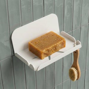 Op de afbeelding: Een wit metalen zeepbakje met een bruin stuk zeep en een houten borstel. Het zeepbakje is bevestigd aan een lichtblauwe betegelde muur. De borstel hangt aan de zijkant.