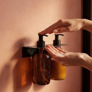 May include: Two amber glass soap dispensers with black pumps mounted on a black metal shelf. The dispensers are against a peach-colored wall. One hand is pressing a pump, and the other is catching the soap.