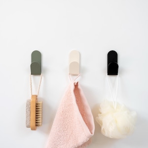 Puede incluir: Tres ganchos de pared en verde oliva, blanco roto y negro, cada uno sosteniendo un artículo de baño diferente. Un cepillo de madera, una toalla rosa y una esponja vegetal amarilla cuelgan de los ganchos, contra una pared blanca.