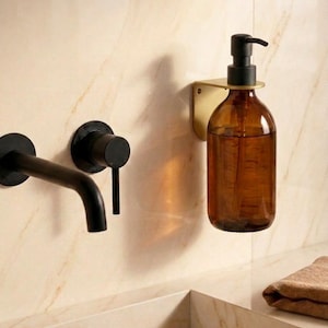 Può includere: Un dispenser di sapone in vetro ambrato montato a parete con pompa nera, sostenuto da una staffa in metallo dorato, posizionato accanto a un rubinetto a parete nero opaco con leva singola su una parete in marmo di colore chiaro.
