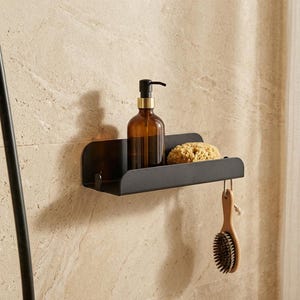 Può includere: Una mensola da parete in metallo nero opaco con base rettangolare e pannello posteriore curvo. La mensola sostiene un flacone con dosatore in vetro ambrato riempito di liquido e una spugna beige naturale. Una spazzola da bagno in legno pende.
