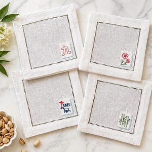 May include: Four square white linen coasters with embroidered details. Each coaster features a different design: a red Chinese character, red flowers, a blue dog with the word "JOKER", and green bamboo. The coasters have a dotted border.