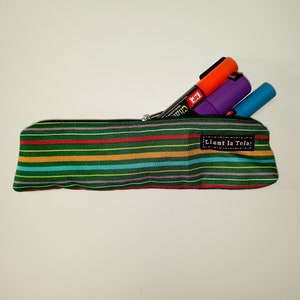 Puede incluir: Un estuche de lápices verde a rayas con cierre de cremallera y una etiqueta que dice "Lian la Tela!" El estuche está lleno de tres marcadores, uno naranja, uno morado y uno azul.