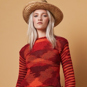 May include: A woman wearing a straw hat and a red and orange knit sweater with long sleeves. The sweater has a unique pattern and is paired with blue denim pants.