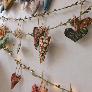 May include: A garland of fabric hearts with floral patterns, hanging on a string with green leaves. The hearts are in various colours, including brown, green, blue, and red.