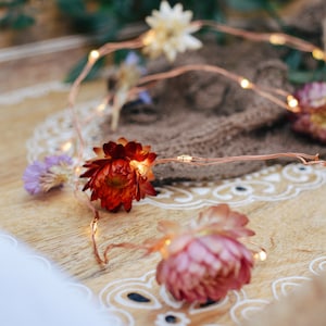 May include: A wooden tray with a white lace doily and a brown burlap cloth. The tray is decorated with dried flowers in shades of red, pink, and purple, and a string of copper wire lights.