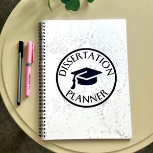 May include: A white spiral bound notebook with a black and white distressed background. The front cover features a black circle with the text "Dissertation Planner" and a black graduation cap icon.
