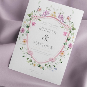Op de afbeelding: Een trouwkaart met een bloemenontwerp. De uitnodiging heeft een roze en wit schildvormig frame met de namen "Jennifer & Matthew" in het midden. De tekst "Save the Date" en "Dinner & Dancing to Follow" zijn ook zichtbaar.