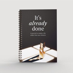Könnte beinhalten: Ein schwarzes Spiralblock-Notizbuch mit dem Text "It's already done" und dem Untertitel "A journal to create a life wilder than your dreams". Das Cover zeigt ein Foto eines Kristallprismas in einem Sonnenstrahl.