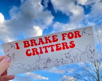 May include: A white bumper sticker with red text reading "I Brake For Critters." The sticker displays line drawings of animals such as a deer, cat, turkey, squirrel, and raccoon, against a cloudy sky.
