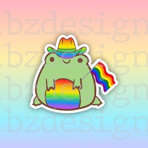 Può includere: Una rana verde con un cappello da cowboy e che tiene una bandiera arcobaleno. La rana è seduta su un cerchio arcobaleno.