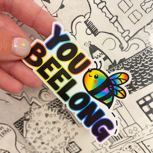 Può includere: Un adesivo arcobaleno con il testo "YOU BELONG" in lettere nere. Accanto al testo c'è un'ape di cartone animato con un corpo arcobaleno.