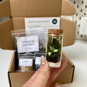 May include: A miniature terrarium kit featuring a small glass bottle with layers of sand, soil, and plants, sealed with a cork. The kit includes a cardboard box with build instructions and packets of substrate and sand. The text "Tiny Terras" is visible.