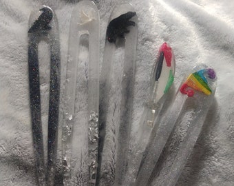 May include: Five clear hair clips with glitter embedded in the plastic. Each clip has a different design: a black bird, a white dove, a black bear, a rainbow, and a multi-colored feather.