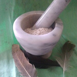 May include: A white marble mortar and pestle with a pile of white rice inside. Two feathers, one brown and one white, are on a green surface.