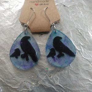 May include: A pair of teardrop-shaped earrings with a blue and purple galaxy-like background. The earrings feature a black silhouette of a crow and a baby crow. The earrings are handmade with love.
