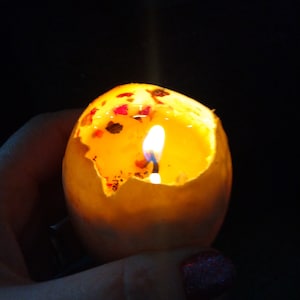 May include: A lit candle made from melted wax inside a cracked eggshell. The candle is yellow and has small red and brown flecks throughout. The eggshell is white.