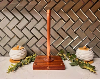 May include: Copper pipe paper towel holder with a wooden base and copper accents. The holder is on a white countertop in front of a gray tile backsplash.
