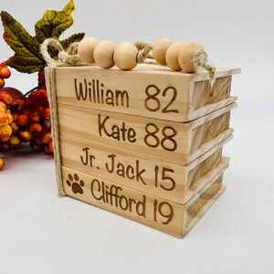 May include: A stack of four wooden blocks with names and numbers engraved on them. The names are William 82, Kate 88, Jr. Jack 15, and Clifford 19. The blocks are tied together with twine and wooden beads.