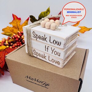 May include: A white wooden book stack with the words "Speak Low If You Speak Love" engraved on the front. The stack is tied with twine and decorated with wooden beads. The stack is sitting on a brown cardboard box with the words "Mit Mit Gift Personalized Gifts and Home Decor" printed on the front.