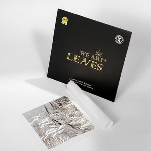 May include: A black box with gold lettering reading "WE ART LEAVES" and a maple leaf logo. Beside it is a sheet of silver leaf and a roll of white paper. The text "Leaves are thin & delicate. Handle with care while opening" is also visible.