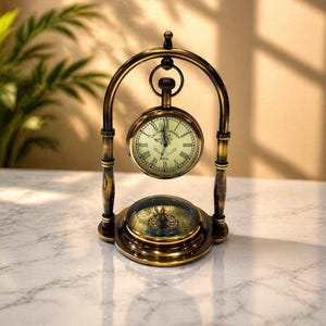 Puede incluir: Un reloj de escritorio antiguo de latón con base de brújula. La esfera del reloj muestra "VICTORIA LONDON 1876" y números romanos. El reloj está suspendido dentro de un marco arqueado decorativo. La brújula es visible debajo.