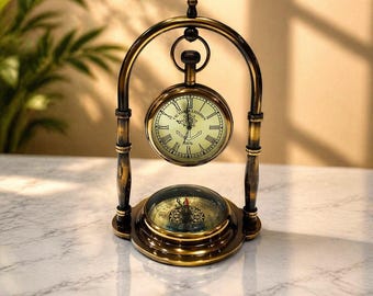 May include: An antique brass desk clock with a compass base. The clock face reads "VICTORIA LONDON 1876" and features Roman numerals. The clock is suspended within a decorative arched frame. The compass is visible below the clock.