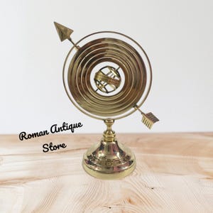 May include: A brass antique armillary sphere with a circular design and arrow accents. The base is round and the sphere has a small globe in the center. The text "Roman Antique Store" is visible on the bottom left.