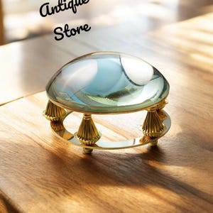 Op de afbeelding: Een heldere glazen vergrootglas met een goudkleurige basis en decoratieve accenten. De tekst "Roman Antique Store" is in zwart schrift boven het vergrootglas geschreven. Het vergrootglas staat op een houten oppervlak.