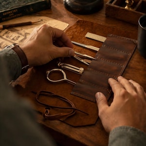 May include: A collection of brass drafting tools, including a compass and dividers, are arranged on a brown leather mat. A brown leather tool roll is open, and a person's hands are visible, suggesting the tools are being organized or used. A book titled "Geometry and Trigonometry" is in the background.