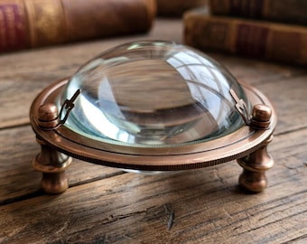 May include: A hemispherical glass magnifying paperweight set within a circular copper-toned metal frame. The frame features a textured, ridged edge and is supported by three small, decorative metal feet, elevating the clear glass dome.