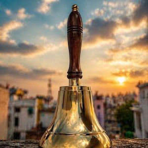 Pu&ograve; includere: Una campana di ottone lucido con un manico in legno scuro e una parte superiore dorata. La campana &egrave; posta contro un tramonto con un paesaggio urbano sullo sfondo. La campana &egrave; alta circa 15 cm.