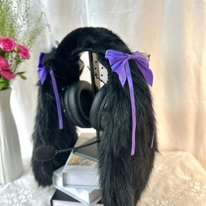 Könnte beinhalten: Schwarze Gaming-Headset-Überzüge aus Kunstfell mit langen, plüschigen Ohrenaufsätzen. Jede Seite ist mit einer leuchtend violetten Satinschleife mit langen, herabhängenden Bändern verziert. Das Headset verfügt über einen schwarzen Mikrofonarm und Ohrpolster.