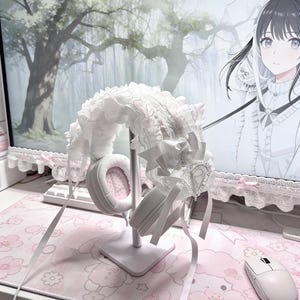May include: White headphones with pink earcups are displayed on a white stand, adorned with a white lace and ribbon headband. The setup is on a pink floral mousepad, creating a soft, feminine aesthetic. A white mouse sits nearby.
