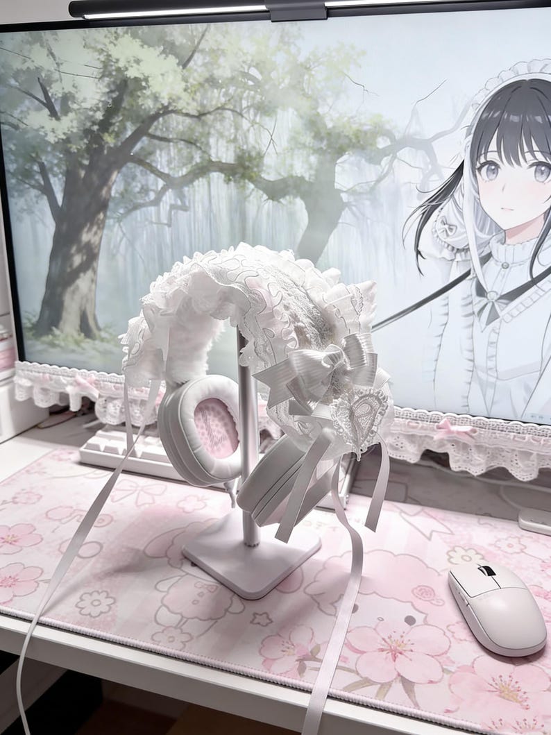 May include: White headphones with pink earcups are displayed on a white stand, adorned with a white lace and ribbon headband. The setup is on a pink floral mousepad, creating a soft, feminine aesthetic. A white mouse sits nearby.