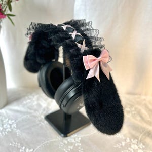 Könnte beinhalten: Schwarze Kopfhörerüberzüge aus Kunstfell mit langen, plüschigen Hasenohren. Der Kopfbügel ist mit schwarzer Spitzenrüsche, einer gekreuzten rosa Bandverzierung und einer kleinen rosa Satinschleife an der Seite eines Ohrs verziert.