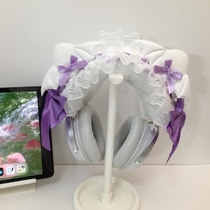 Puede incluir: Un soporte blanco para auriculares con una diadema decorativa de alas de murciélago con lazos morados y ribete de encaje. Los auriculares grises cuelgan del soporte. Una tableta es visible en el fondo.