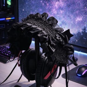 May include: Black headset with red accents on a black stand, adorned with a black, ornate headpiece featuring lace and ribbons. The headpiece is styled with a gothic aesthetic. The background includes a computer monitor displaying a galaxy.