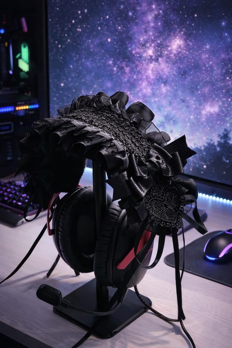 May include: Black headset with red accents on a black stand, adorned with a black, ornate headpiece featuring lace and ribbons. The headpiece is styled with a gothic aesthetic. The background includes a computer monitor displaying a galaxy.