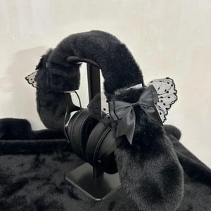 May include: Black headphones with faux fur ear covers and headband. The ear covers have a fluffy texture and are decorated with black bows and lace detailing. The headphones are displayed on a black stand against a neutral background.