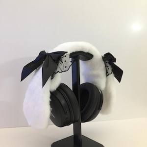 Puede incluir: Auriculares negros sobre la oreja en un soporte negro, adornados con una funda de piel sintética blanca. La funda presenta lazos negros y detalles de encaje. Los auriculares se muestran sobre un fondo blanco.