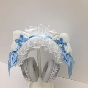 Puede incluir: Accesorio de auriculares blanco y azul con un diseño decorativo. El accesorio presenta tela blanca, ribete de encaje y lazos azul claro. Los auriculares son plateados y grises, apoyados en un soporte blanco.