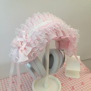 Puede incluir: Un gorro rosa y blanco con ribete de encaje y un lazo. El gorro se exhibe en un soporte blanco con un par de auriculares blancos. El fondo es un patrón de cuadros rosas y blancos.