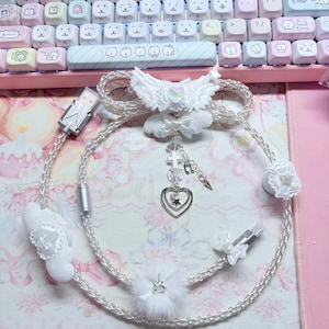 Puede incluir: Un cable de carga trenzado blanco con detalles decorativos que incluyen un par de alas, un pompón esponjoso y un dije colgante con una cruz, una nota musical y un corazón doble. El cable incluye protectores en forma de lazo.