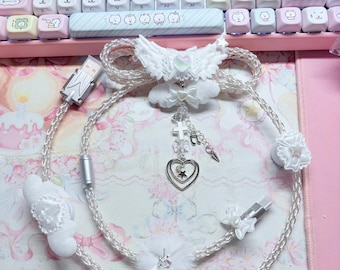 Puede incluir: Un cable de carga trenzado blanco con detalles decorativos que incluyen un par de alas, un pompón esponjoso y un dije colgante con una cruz, una nota musical y un corazón doble. El cable incluye protectores en forma de lazo.