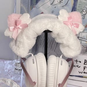 Könnte beinhalten: Weiße Kopfhörer mit einem flauschigen, weißen Stirnband mit rosa Akzenten und Schleifen. Das Stirnband hat rosa ohrenförmige Details. Die Kopfhörer haben ein rosa Band und werden auf einem schwarzen Ständer präsentiert.